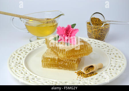 Favo di miele, fiore rosa sottili su piastra di porcellana con cucchiaio vintage di polline accanto e ciotole di miele e polline in background Foto Stock