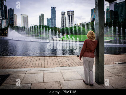 Signora bionda appoggiata contro un post guardare il display di acqua nella parte anteriore della Petronas Twin Towers, Kuala Lumpur, Malesia Foto Stock