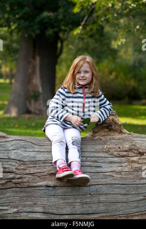 Una giovane ragazza con i capelli di zenzero è sorridente ampiamente come lei si siede in alto su un albero caduto trunk. Foto Stock