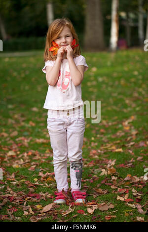 Una giovane ragazza con i capelli di zenzero contiene due belle foglie di autunno contro il suo volto come lei gioca su una soleggiata giornata autunnale. Foto Stock