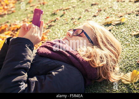 Bionda caucasica ragazza adolescente in giacca nera che stabilisce nel parco autunnali e utilizzando il cellulare per la messaggistica in rete sociale Foto Stock
