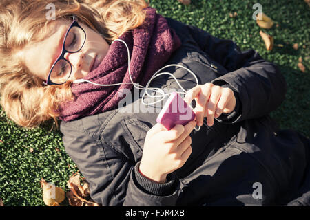Bionda caucasica ragazza adolescente in giacca nera che stabilisce nel parco autunnali e utilizza lo smartphone Foto Stock