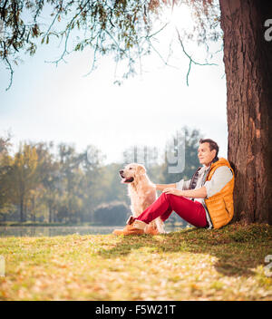 Giovane uomo seduto da un albero in posizione di parcheggio con il suo cane su una soleggiata giornata autunnale shot con inclinazione e spostamento lente Foto Stock