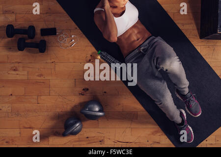 Vista superiore della donna muscolare facendo sit ups. Femmina giacente sul tappeto di esercizio facendo allenamento dello stomaco con equipaggiamenti da palestra in piano. Foto Stock