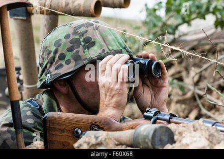 La seconda guerra mondiale la rievocazione. Funzionario tedesco indossando il camuffamento camiciotto e casco, in trincea guardando attraverso il binocolo, stick bombe accanto a lui Foto Stock