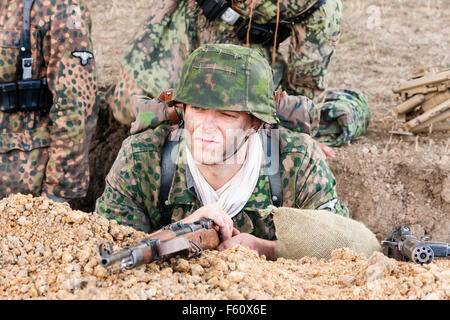 La seconda guerra mondiale la rievocazione. Soldato tedesco tenendo il coperchio in trincea, in guardia, tenendo fucile. Indossando il camuffamento camiciotto e casco. Foto Stock