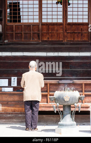 Giappone, Kyoto, Seiryo-ji. Vista da dietro del senior uomo in piedi di fronte a temple hall pregando accanto al fumo di incenso bruciatore. Foto Stock