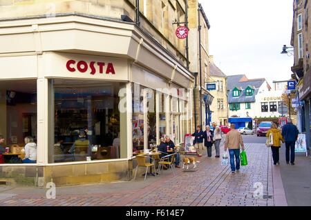 La gente seduta al di fuori e oltrepassando caffè costa all'angolo del pasto e mercato Fore Street, Hexham, Northumberland, Regno Unito. Foto Stock