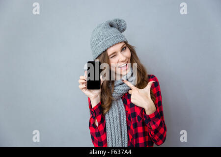 Ritratto di una donna allegra winking e puntare il dito sullo sbozzato lo schermo dello smartphone su uno sfondo grigio Foto Stock