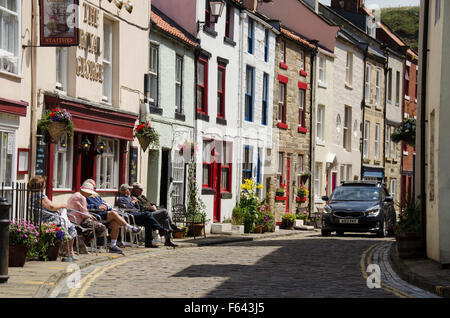 Auto giri l'angolo come persone sedersi al sole e rilassarsi al di fuori del pub - acciottolato stretto High Street, pittoresco villaggio di Staithes, North Yorkshire, Regno Unito. Foto Stock