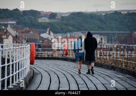 Coppia giovane a piedi (il suo braccio attorno alla sua vita) in una serata estiva, lungo il Molo Ovest di estensione. La cittadina di Whitby, North Yorkshire, GB, oltre. Foto Stock