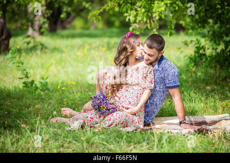 Felice coppia giovane aspetta baby, donna incinta baciare il marito, seduti su erba verde Foto Stock