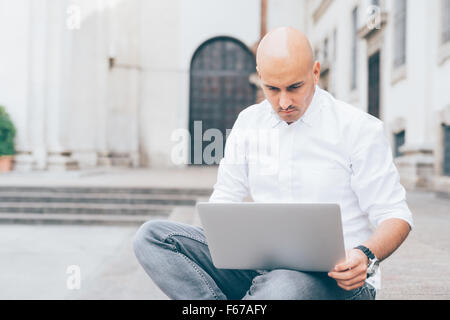 Giovani caucasici bello bald business uomo seduto su una scala utilizzando un laptop appoggiato sulle ginocchia, guardando verso il basso lo schermo, pensieroso - lavoro, meditata, concetto di occupato Foto Stock