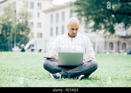 Giovani caucasici bello bald business uomo seduto in un parco della città utilizzando un laptop appoggiato sulle ginocchia, guardando verso il basso lo schermo, pensieroso - lavoro, meditata, concetto di occupato Foto Stock