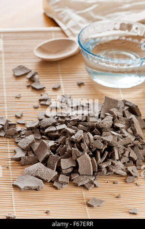 Ghassoul lava pezzi di argilla, essiccato chip rovesciato, ciotola di vetro con acqua e il cucchiaio di legno sul tappetino, sbriciolato Rhassoul marocchino... Foto Stock