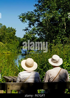 Coppia di anziani che indossa cappello da sole seduto su una panca in legno godendo di una calda giornata estiva in prossimità di un lago Foto Stock