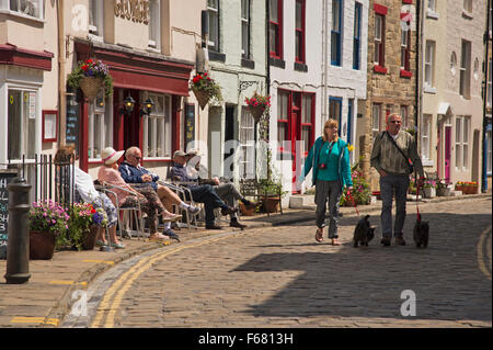 Persone, rilassante seduta al di fuori di un pub o a piedi lungo la soleggiata acciottolate High Street - tradizionale villaggio di pescatori di Staithes, North Yorkshire, Regno Unito. Foto Stock