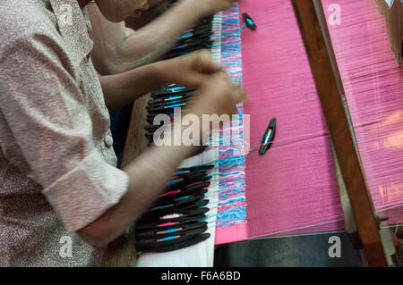 Le mani di una donna di tessitura utilizzando navette con bobine di filo su di un telaio di tessitura. Parzialmente tessuto tessuto di seta visibile. Mandalay, Myanmar. Foto Stock