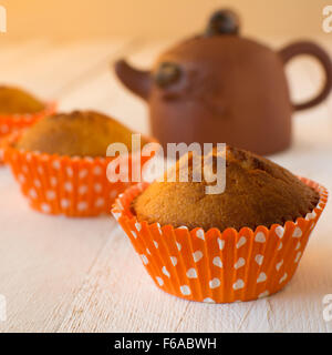 Muffin in carta arancione cupcake titolare con il bianco a pois marrone e teiera ceramica su un bianco sullo sfondo di legno, quadrato Foto Stock