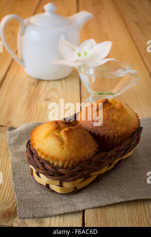 Due muffin in un piccolo cesto in vimini su tela, fiore bianco e teiera in porcellana su una tavola di legno, fuoco selettivo Foto Stock