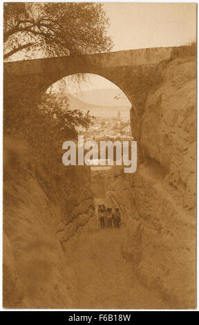 Questa cartolina fotografica vintage Real Photo (RPPC) presenta uomini che si trovano sotto un arco di pietra, con acquedotti e archi sullo sfondo, che riflettono le caratteristiche architettoniche storiche comunemente presenti nelle città europee. Foto Stock