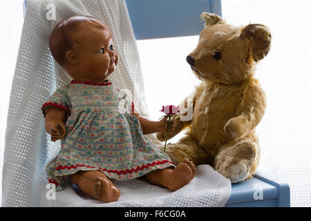 Vintage bambola di ceramica apparentemente offre una nuova red rose di un orso di peluche su una coltre bianca su una sedia blu. Sfondo bianco Foto Stock