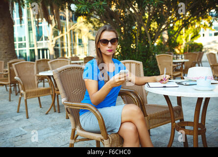 Candida immagine di un giovane piuttosto moderno blogger utilizza lo smartphone e prende appunti in un notebook in un cafe Foto Stock