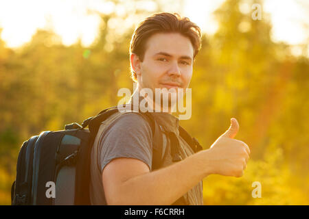 Uomo giovani turisti con zaino che mostra Thumbs up handsign. Rosso la luce del tramonto. Foto Stock
