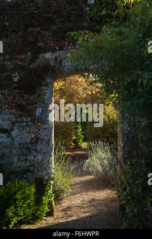 Un arco in pietra del Cotswold adagiato in un alto muro di mattoni retroilluminato dal caldo sole di ottobre nel giardino recintato, i giardini di Rousham House, Oxfordshire, Inghilterra. Foto Stock