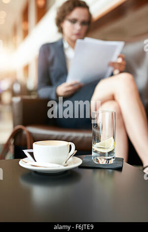 Tazza di caffè e un bicchiere di acqua sul tavolo del bar con imprenditrice seduta in background la lettura dei documenti. Foto Stock