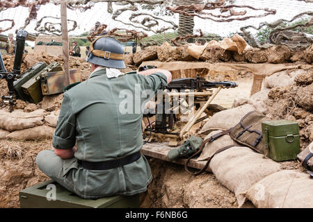 La seconda guerra mondiale la rievocazione. Soldato tedesco seduto, con Torna al visualizzatore, in trincea sandbagged manning MG42 mitragliatrice sotto camuffamento di netting. Foto Stock