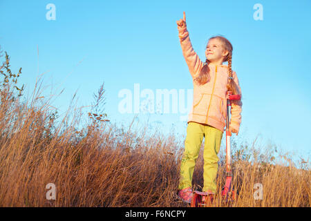 Bambina abiti brillanti con gli scooter mostra un dito verso il cielo per la natura Foto Stock