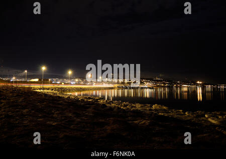 Lungo litorale innevato con luce riflettendo sulla superficie con tromsoe città illuminazione notturna in background Foto Stock
