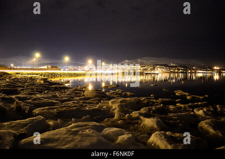 Lungo litorale innevato con luce riflettendo sulla superficie con tromsoe città illuminazione notturna in background Foto Stock
