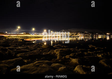 Lungo litorale innevato con luce riflettendo sulla superficie con tromsoe città illuminazione notturna in background Foto Stock