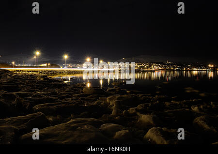 Lungo litorale innevato con luce riflettendo sulla superficie con tromsoe città illuminazione notturna in background Foto Stock