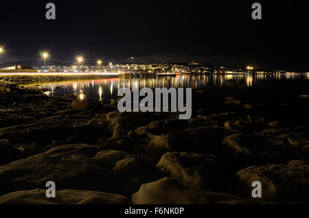 Lungo litorale innevato con luce riflettendo sulla superficie con tromsoe città illuminazione notturna in background Foto Stock