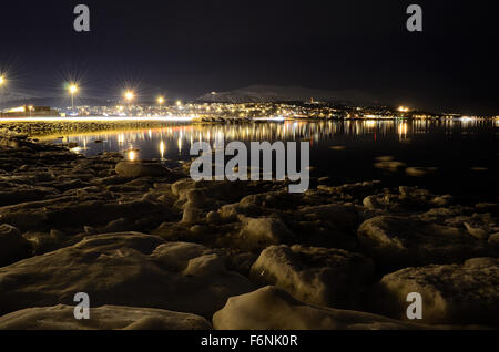 Lungo litorale innevato con luce riflettendo sulla superficie con tromsoe città illuminazione notturna in background Foto Stock