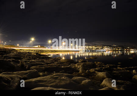 Lungo litorale innevato con luce riflettendo sulla superficie con tromsoe città illuminazione notturna in background Foto Stock