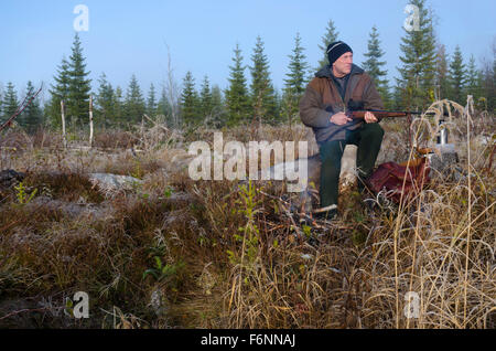 Moose hunter seduti su un moncone con un po' fuoco davanti tenendo il suo fucile rivolto verso sinistra. Foto Stock