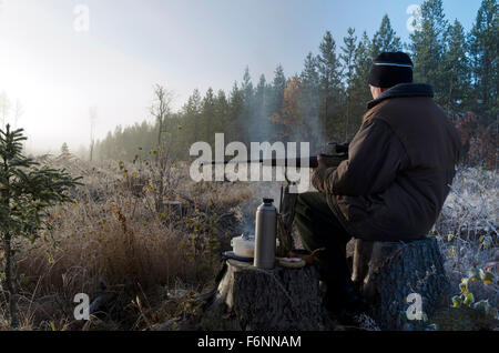 Moose hunter seduti su un moncone con un po' fumoso fuoco davanti tenendo un fucile e con un sandwich e un thermos in attesa. Foto Stock