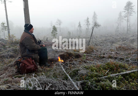 Foto da destra di un cacciatore di alci seduto su un moncone di prima mattina con un po' fuoco davanti tenendo un fucile, immagine Foto Stock