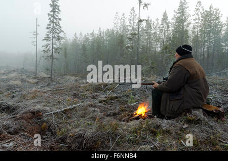 Foto da sinistra di un cacciatore di alci seduto su un moncone di prima mattina con un po' di fuoco davanti tenendo un fucile. Foto Stock