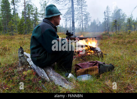 Moose cacciatore con speciale panno homespun seduti su un moncone con un po' fuoco davanti tenendo un fucile. Foto Stock