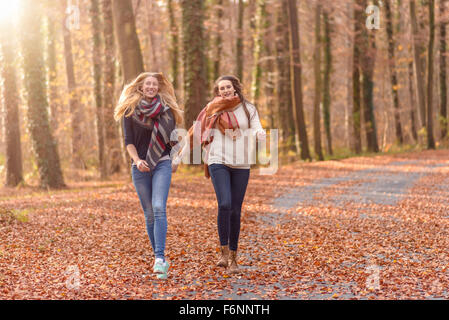 Due donne gioiosa esecuzione di mano in mano attraverso una boscosa del parco in autunno con le foglie colorate che giace a terra in un sano lifesty Foto Stock