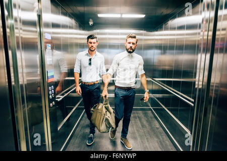 Due giovani bello nero e capelli biondi imprenditore moderno che esce da un ascensore, una holding di un sacchetto, entrambi affacciati a destra - business, lavorando, concetto di successo Foto Stock