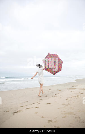 Donna che cammina nel vento con ombrellone in spiaggia Foto Stock