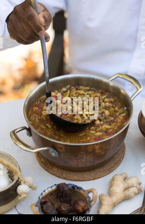 Un uomo è ladling harira in un recipiente esterno in una collezione autunno ambiente picnic. La zuppa viene servita con maggiorana fresca e coriandolo su t Foto Stock