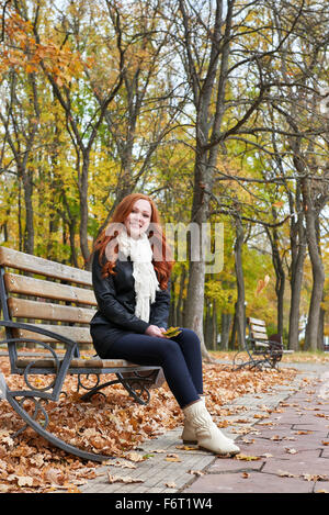 Redhead ragazza seduta su una panchina nel parco cittadino, stagione autunnale Foto Stock