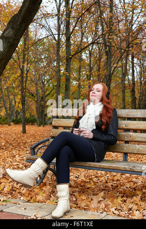 Redhead girl ascoltare musica su cuffie in city park, caduta stagione Foto Stock
